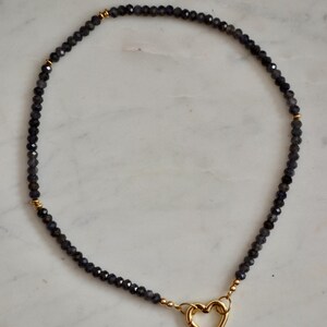 Op de afbeelding: Een donkergrijze kralenketting met een gouden hartvormige sluiting. De ketting bestaat uit kleine, facetgeslepen kralen en gouden accenten. De hartsluiting is gecentreerd, wat zorgt voor een delicaat en elegant ontwerp.