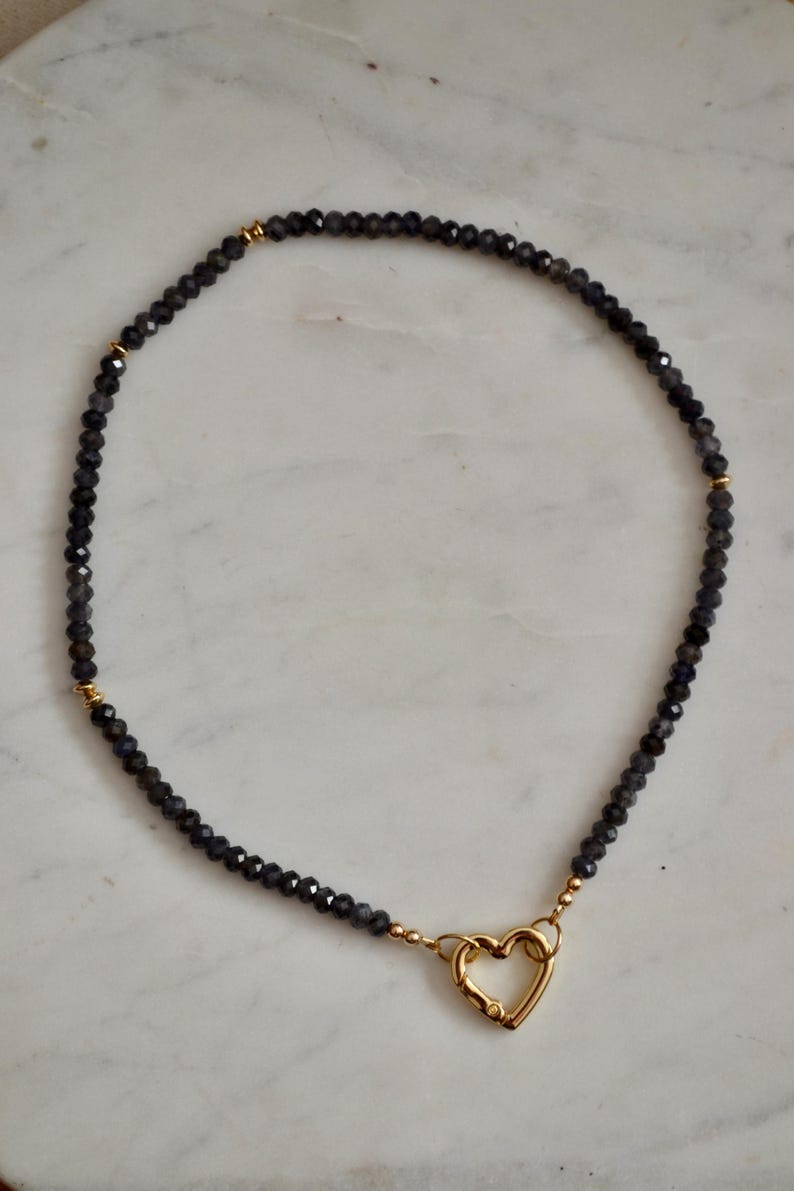 Op de afbeelding: Een donkergrijze kralenketting met een gouden hartvormige sluiting. De ketting bestaat uit kleine, facetgeslepen kralen en gouden accenten. De hartsluiting is gecentreerd, wat zorgt voor een delicaat en elegant ontwerp.