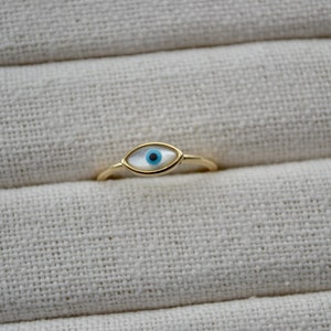 May include: A gold-colored ring featuring an eye-shaped design. The eye has a white background with a blue iris and a black pupil. The ring is set against a textured, off-white fabric.