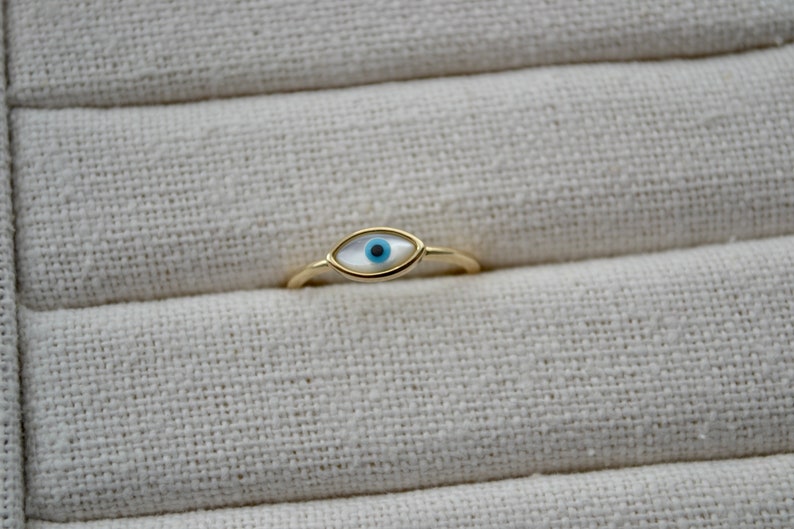 May include: A gold-colored ring featuring an eye-shaped design. The eye has a white background with a blue iris and a black pupil. The ring is set against a textured, off-white fabric.