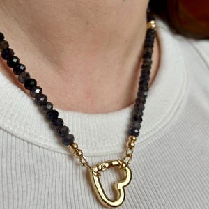 Op de afbeelding: Een ketting met een gouden hartvormige sluiting en donkere, facetgeslepen kralen. De ketting heeft gouden accenten en wordt gedragen op een wit geribbeld shirt. De kralen lijken een donkere, iriserende kleur te hebben.