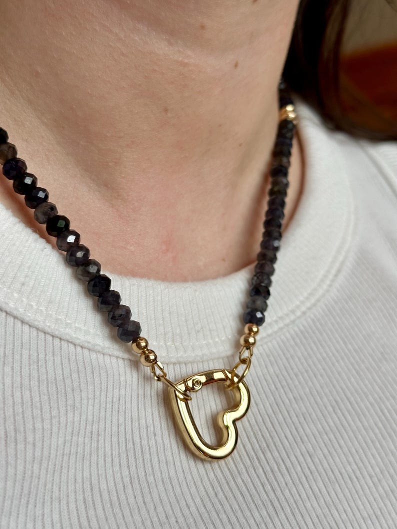 Op de afbeelding: Een ketting met een gouden hartvormige sluiting en donkere, facetgeslepen kralen. De ketting heeft gouden accenten en wordt gedragen op een wit geribbeld shirt. De kralen lijken een donkere, iriserende kleur te hebben.