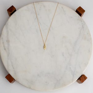 May include: A gold pendant necklace with a teardrop-shaped charm. The necklace is displayed on a round, white marble surface with wooden feet. The pendant has a small, oval design.