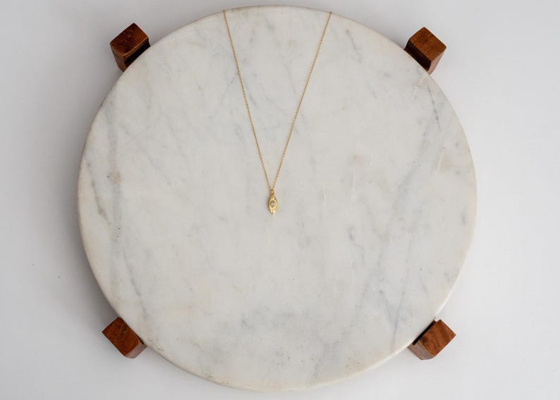 May include: A gold pendant necklace with a teardrop-shaped charm. The necklace is displayed on a round, white marble surface with wooden feet. The pendant has a small, oval design.