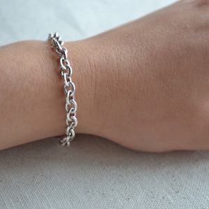 May include: A silver-colored chain bracelet with a series of oval links is worn on a wrist. The bracelet is made of a shiny metal and is secured with a clasp. The skin tone is light, and the background is a neutral color.