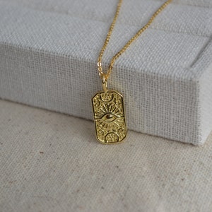 May include: Gold necklace with a rectangular pendant featuring an eye with sun rays and moon phases. The pendant has a detailed, ornate design.