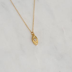 May include: A gold-coloured necklace featuring an oval pendant with a central clear stone and decorative detailing. The fine chain and pendant are set against a white marble surface. The necklace is a piece of jewellery.