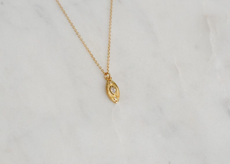 May include: A gold-coloured necklace featuring an oval pendant with a central clear stone and decorative detailing. The fine chain and pendant are set against a white marble surface. The necklace is a piece of jewellery.