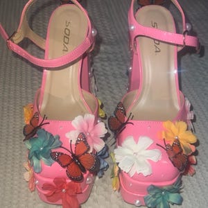 May include: Bright pink platform heels with ankle straps, adorned with colorful fabric flowers, butterflies, and faux pearls. The heels have the brand name "SODA" printed on the insole.