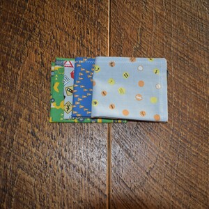 May include: Four fabric squares, stacked on top of each other. The top square is light blue with orange and yellow polka dots. The second square is blue with a pattern of yellow cones. The third square is blue with a pattern of yellow and white traffic signs. The bottom square is green with a pattern of yellow construction equipment.