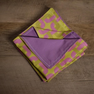 Puede incluir: Servilleta de tela doblada con un estampado geométrico en verde lima y rosa, con un interior liso lavanda. La servilleta está colocada sobre una superficie de madera.