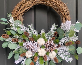 May include: A rustic grapevine wreath adorned with faux frosted pine, eucalyptus leaves, cotton bolls, and pinecones. The wreath is a festive decoration for the holiday season, adding a touch of natural beauty to any door or wall.