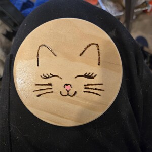 May include: A circular light-colored wooden coaster featuring a wood-burned cat face design. The cat features two pointed ears, closed eyes with long eyelashes, three whiskers on each side, and a small, hand-painted pink nose in the center.