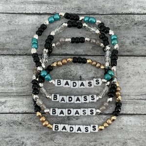 Puede incluir: Cuatro pulseras de cuentas con la palabra "BADASS" escrita en cuentas de letras blancas. Las pulseras están superpuestas y presentan cuentas negras, plateadas, doradas y turquesas.