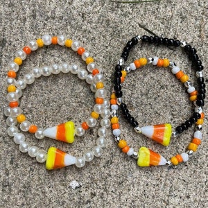 Può includere: Due braccialetti di perline con ciondoli a forma di mais dolce. Un braccialetto è bianco con perline arancioni e gialle, l'altro è nero con perline bianche e arancioni.