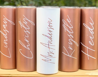 May include: Five personalized tumblers with names written in white script. The tumblers are rose gold in color, except for one which is white.