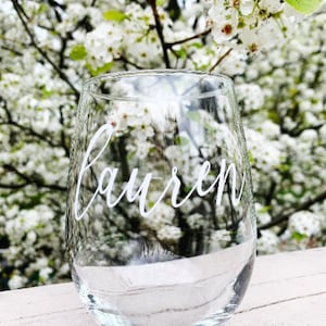 May include: A clear glass wine glass with the name "Lauren" written in white lettering on the side. The glass is sitting on a white wooden surface with a blurry background of white flowers.