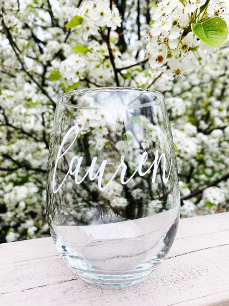 May include: A clear glass wine glass with the name "Lauren" written in white lettering on the side. The glass is sitting on a white wooden surface with a blurry background of white flowers.