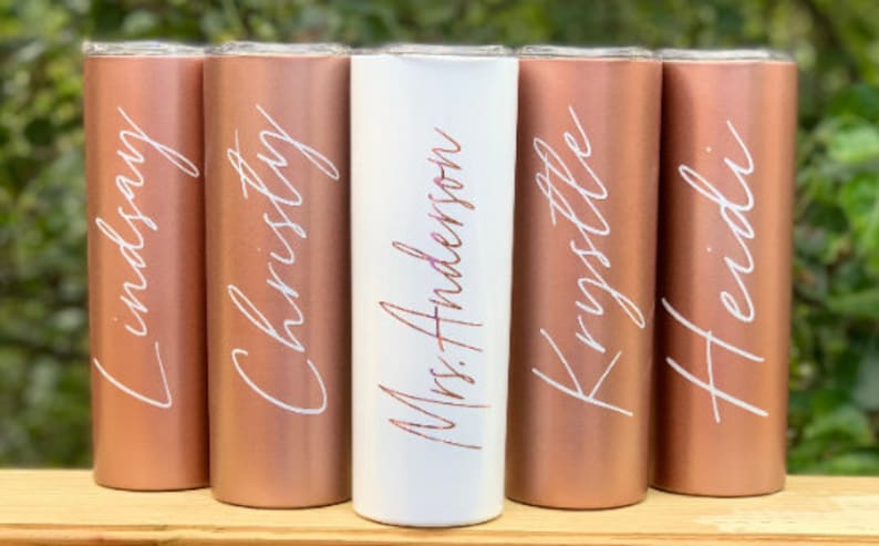 May include: Five personalized tumblers with rose gold and white finishes. The tumblers are labeled with the names "Lindsay", "Christy", "Mrs. Anderson", "Krystle", and "Heidi".