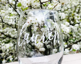 May include: A clear stemless wine glass with the name "Lauren" in white script. The glass is set against a backdrop of white blossoms and a light-colored wooden surface.