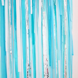 May include: A backdrop made of blue, white, and silver streamers. The streamers are hanging vertically and are all different lengths. The backdrop is perfect for a party or event.