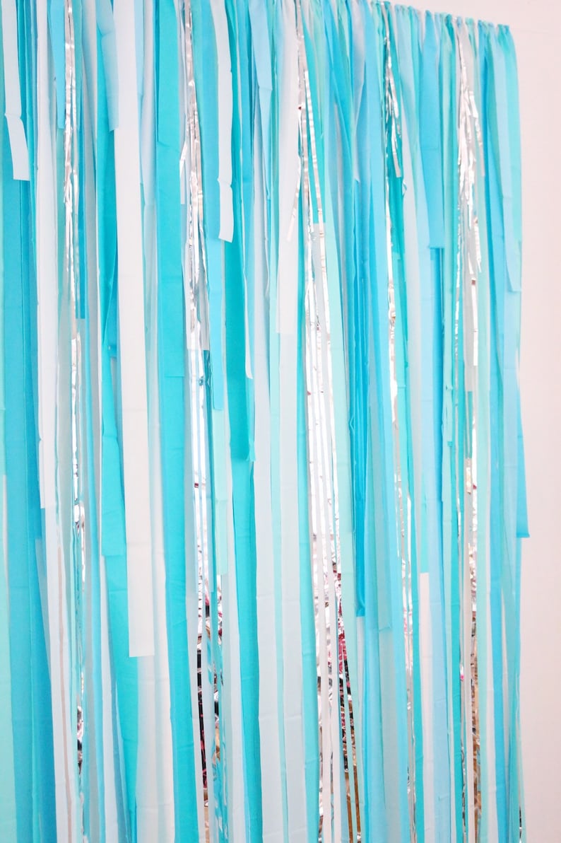 May include: A backdrop made of blue, white, and silver streamers. The streamers are hanging vertically and are all different lengths. The backdrop is perfect for a party or event.