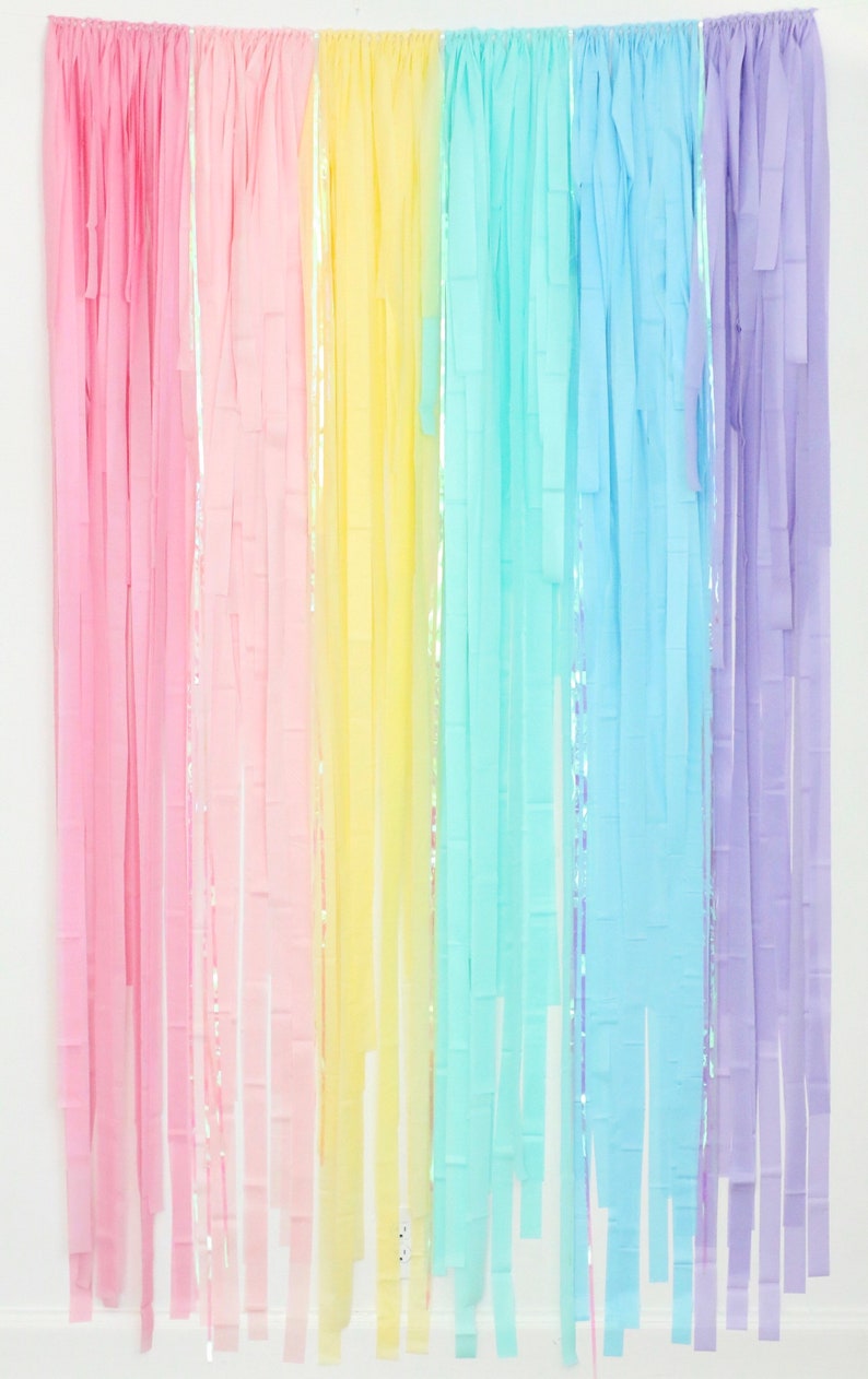May include: Rainbow colored tissue paper streamers hanging from a white wall. The streamers are in shades of pink, yellow, blue, and purple.