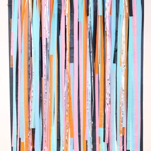 May include: A backdrop made of colorful streamers in black, orange, pink, and light blue. The streamers are hanging from a string and are arranged in a vertical pattern.