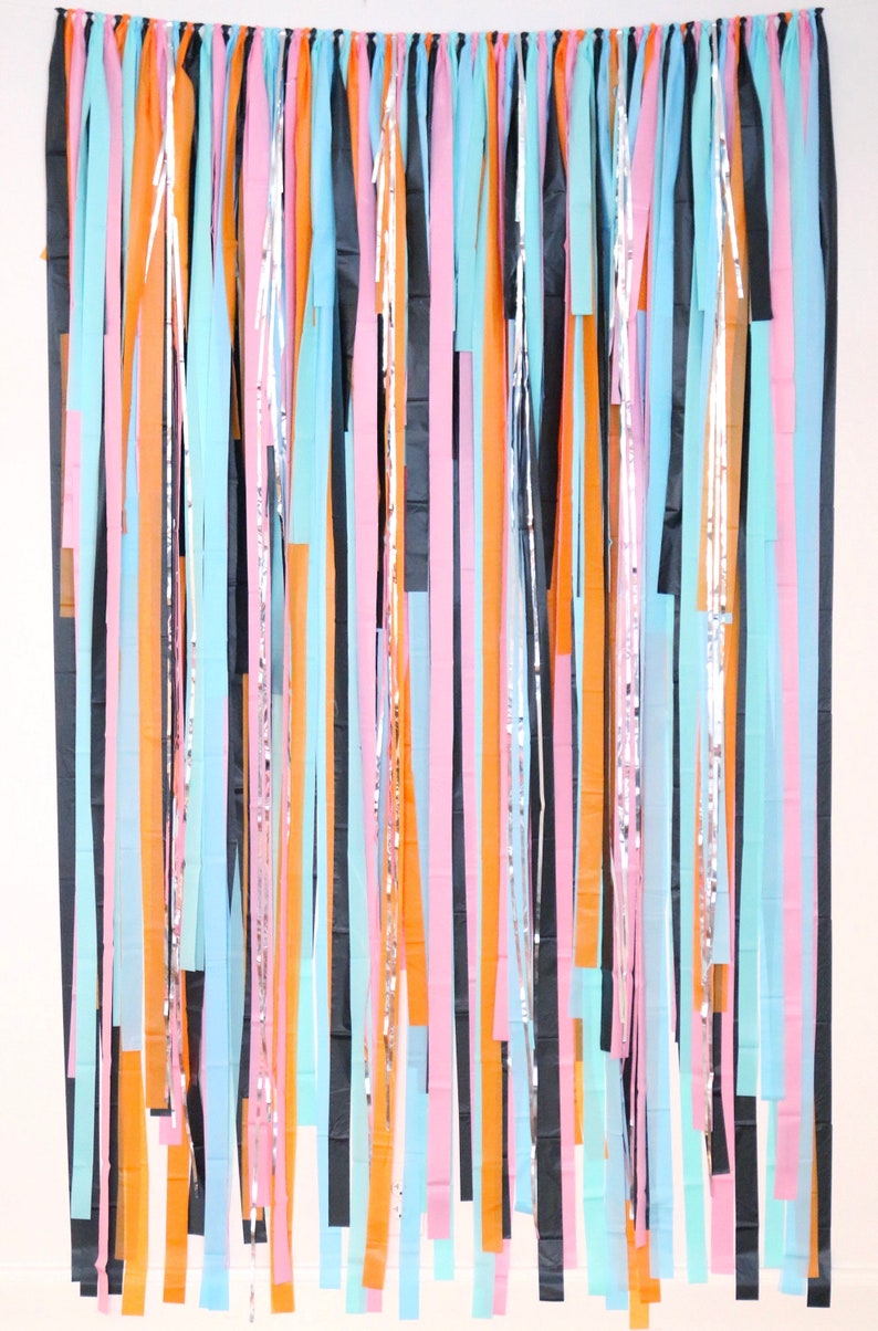 May include: A backdrop made of colorful streamers in black, orange, pink, and light blue. The streamers are hanging from a string and are arranged in a vertical pattern.
