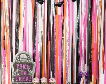 May include: A Halloween display featuring a backdrop of pink, orange, black, and silver streamers. A shelf displays cupcakes, a tombstone sign reading "Trick or Treat", a spiderweb bowl, and skull decorations. A string of felt bats hangs above.