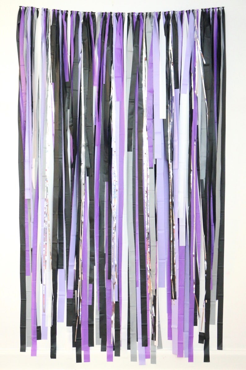 May include: A backdrop made of purple, black, and white streamers hanging from a string. The streamers are all different lengths and are arranged in a random pattern.