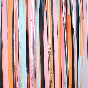 May include: A colorful backdrop made of strips of paper and metallic foil. The strips are in various colors, including orange, pink, blue, black, and silver. The strips are hanging from a ceiling or wall, creating a festive and celebratory atmosphere.