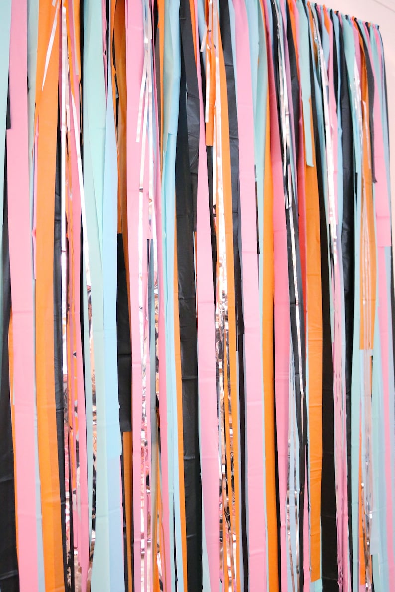 May include: A colorful backdrop made of strips of paper and metallic foil. The strips are in various colors, including orange, pink, blue, black, and silver. The strips are hanging from a ceiling or wall, creating a festive and celebratory atmosphere.
