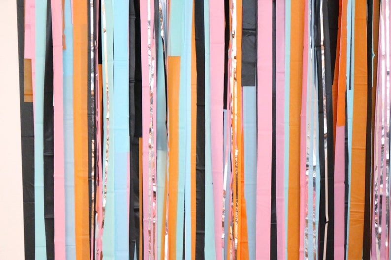 May include: A colorful backdrop of streamers in various colors, including black, orange, pink, blue, and silver. The streamers are hanging vertically and create a festive and celebratory atmosphere.