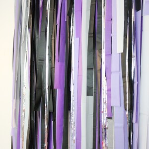 May include: A backdrop made of purple, black, silver, and white streamers hanging from the ceiling. The streamers are all different lengths and create a festive and whimsical look.