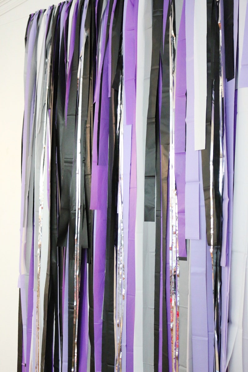 May include: A backdrop made of purple, black, silver, and white streamers hanging from the ceiling. The streamers are all different lengths and create a festive and whimsical look.