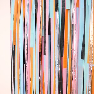 May include: A colorful backdrop made of strips of paper in various colors, including pink, orange, blue, black, and silver. The strips are hanging vertically and create a festive and whimsical look.