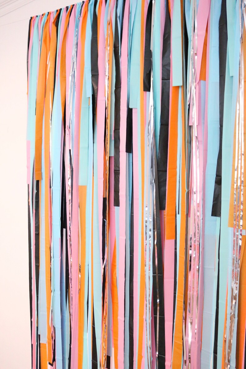 May include: A colorful backdrop made of strips of paper in various colors, including pink, orange, blue, black, and silver. The strips are hanging vertically and create a festive and whimsical look.