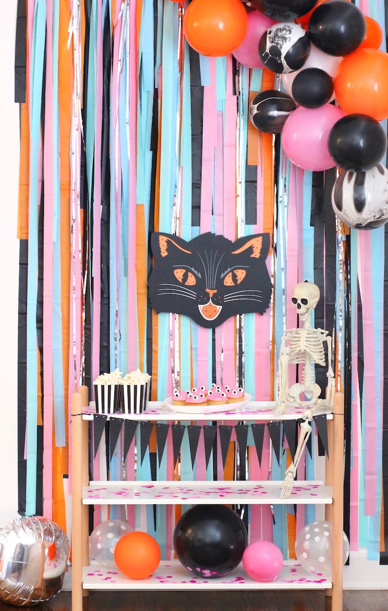 May include: A Halloween party backdrop with black, orange, pink, and blue streamers. A black cat head cutout is hanging on the wall. A white shelf with two tiers is decorated with black and white striped popcorn containers, pink cupcakes with googly eyes, and a skeleton figurine. The shelf is decorated with black triangle flags and pink confetti.