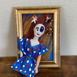Puede incluir: Una muñeca blanca con un ojo azul y un ojo negro, que lleva un vestido de lunares azules y blancos con cuentas rojas alrededor del cuello. La muñeca está en un marco dorado con un fondo pintado y cuatro pequeñas muñecas colgando del marco.