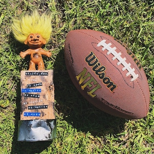 以下が含まれることがあります： 黄色の髪の毛のトロール人形が、白いステッチと「Wilson NFL」の文字が印刷された茶色の革製のフットボールの隣に立っています。フットボールは緑の芝生の上にあります。