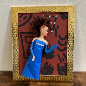 Könnte beinhalten: Eine Puppe in einem blauen Kleid mit Strassdetails ist in einem goldfarbenen, strukturierten Rahmen gerahmt. Der Hintergrund ist weinrot mit schwarzen abstrakten Formen und einigen Strasssteinen. Die Puppe hat braunes Haar.