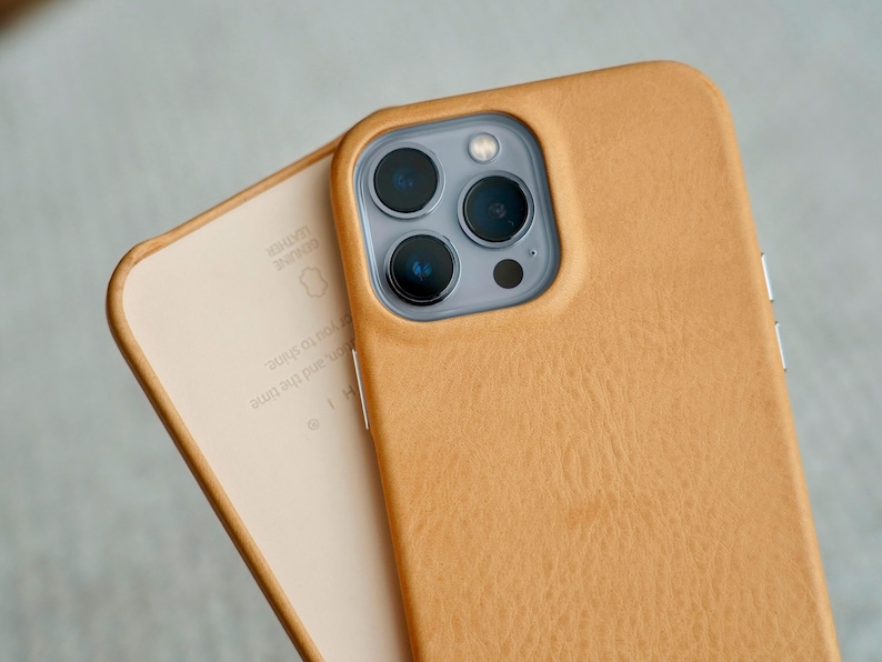 May include: Close-up of two tan leather phone cases. One case is on top of the other, revealing the interior of the bottom case. The top case has a camera module with three lenses. The leather has a textured appearance.