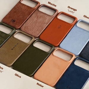 May include: A collection of leather phone cases in various colors, including brown, orange, blue, and green. Each case has a cutout for the camera and is labeled with a color name. Some cases have text that reads "Mocha", "Orange", "Ghost Blue", "Old Oliva", "Oliva", and "Natur".