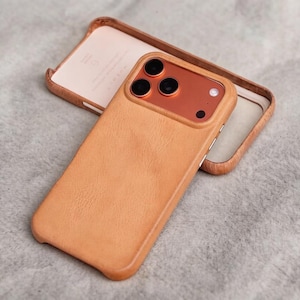 May include: A tan leather phone case with a wooden frame, and a separate wooden frame. The phone case has a textured surface and a copper-colored camera module with three black lenses. The wooden frame is light brown.