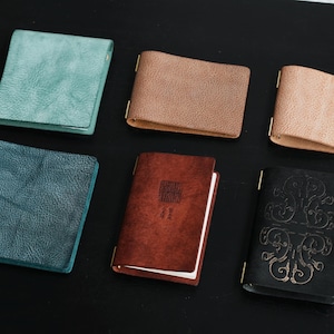 May include: An assortment of leather journals and wallets in various colors, including teal, brown, and black. The journals have gold-colored metal clasps and some feature embossed designs or text. The items are arranged on a dark surface.