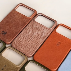 Pode incluir: Uma coleção de capas de telefone de couro em várias cores, incluindo marrom, bege e laranja. Cada capa apresenta um recorte retangular para a câmera e uma superfície texturizada. Algumas capas têm um logotipo em relevo.