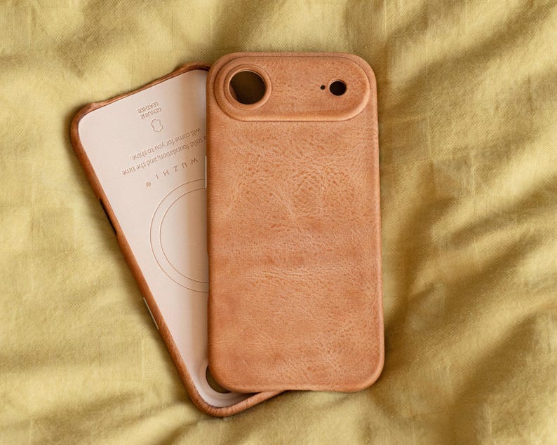 May include: A light brown leather phone case with a matching interior. The case has a textured surface and a cutout for the camera. The interior has the text "WUZHI" and other text. The case is lying on a yellow surface.