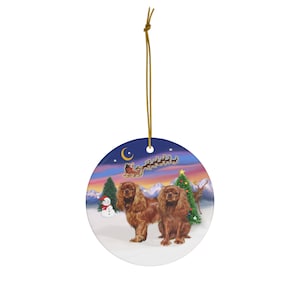 May include: A round Christmas ornament featuring two Cavalier King Charles Spaniels in a winter scene. The ornament has a gold string for hanging. Santa's sleigh and reindeer are in the sky above the dogs.