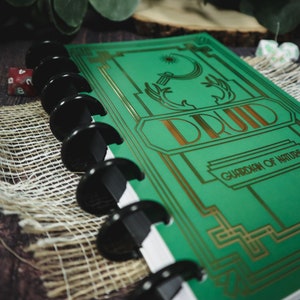 Op de afbeelding: Een groen en gouden spiraalnotitieblok met het woord "DRUID" in gouden letters op de voorzijde. De omslag heeft ook een gouden ontwerp en de tekst "Bewaker van de natuur".